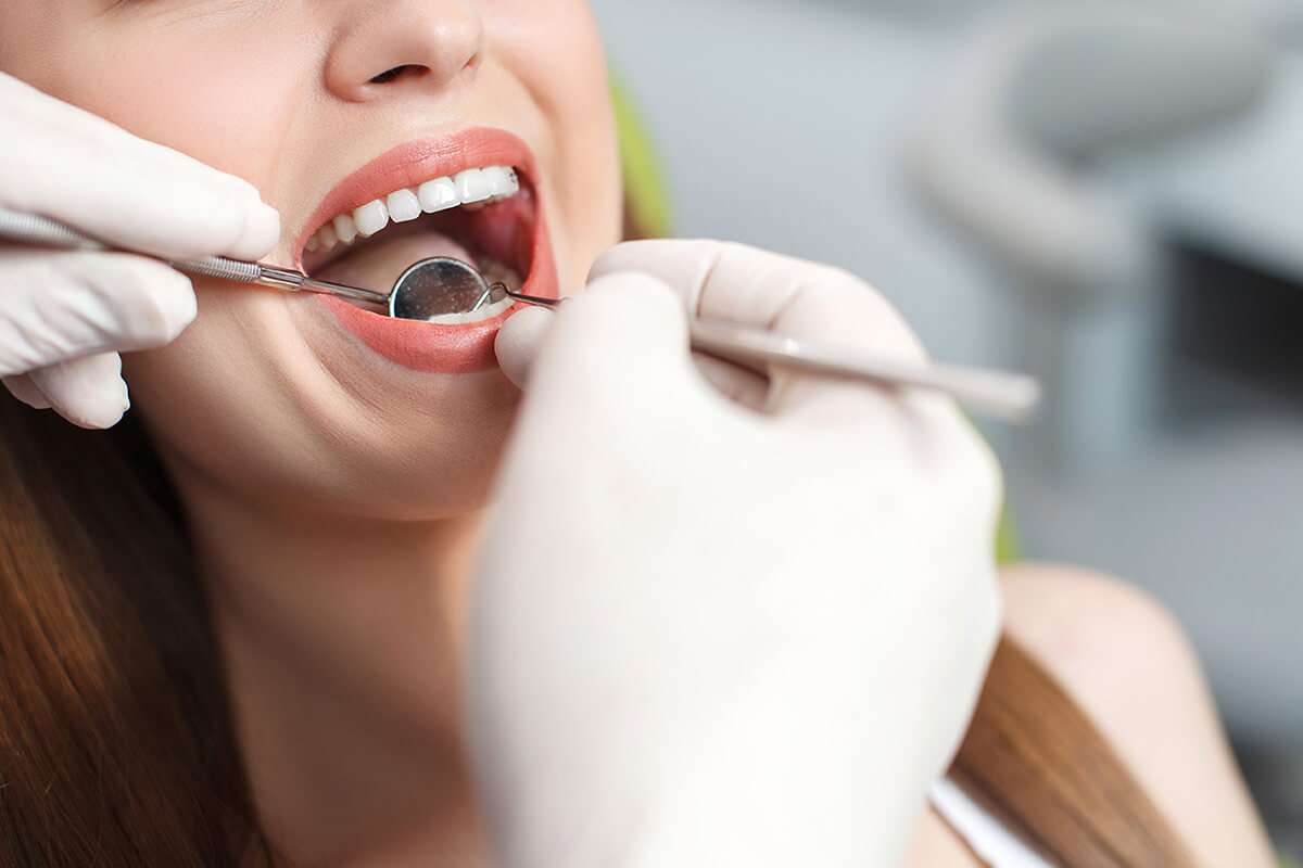 A dentist wearing gloves examines a patients teeth using a dental mirror and probe. The patient has their mouth open, showing clean, white teeth.