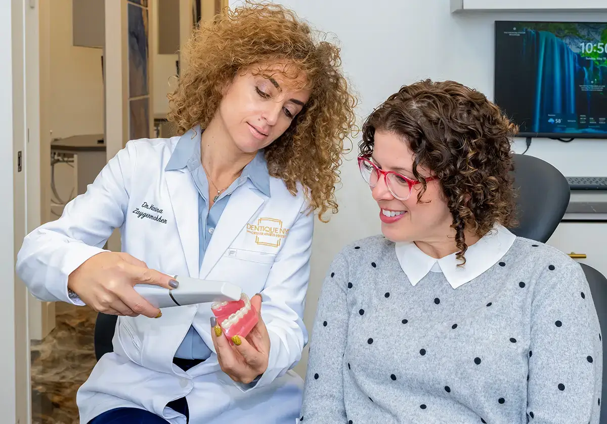 A dentist in a white coat demonstrates a dental procedure using a dental model and a tool to a seated patient with curly hair and red glasses, who is smiling and wearing a gray polka dot sweater, in a clinic setting.