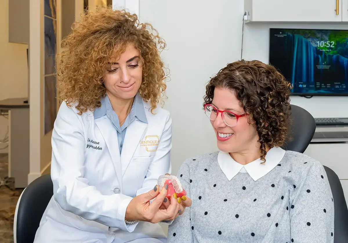 A dental professional with curly hair and a white coat is showing a dental model to a smiling patient with curly hair and red glasses, who is seated and wearing a white blouse with black polka dots. They appear to be in a dental office.