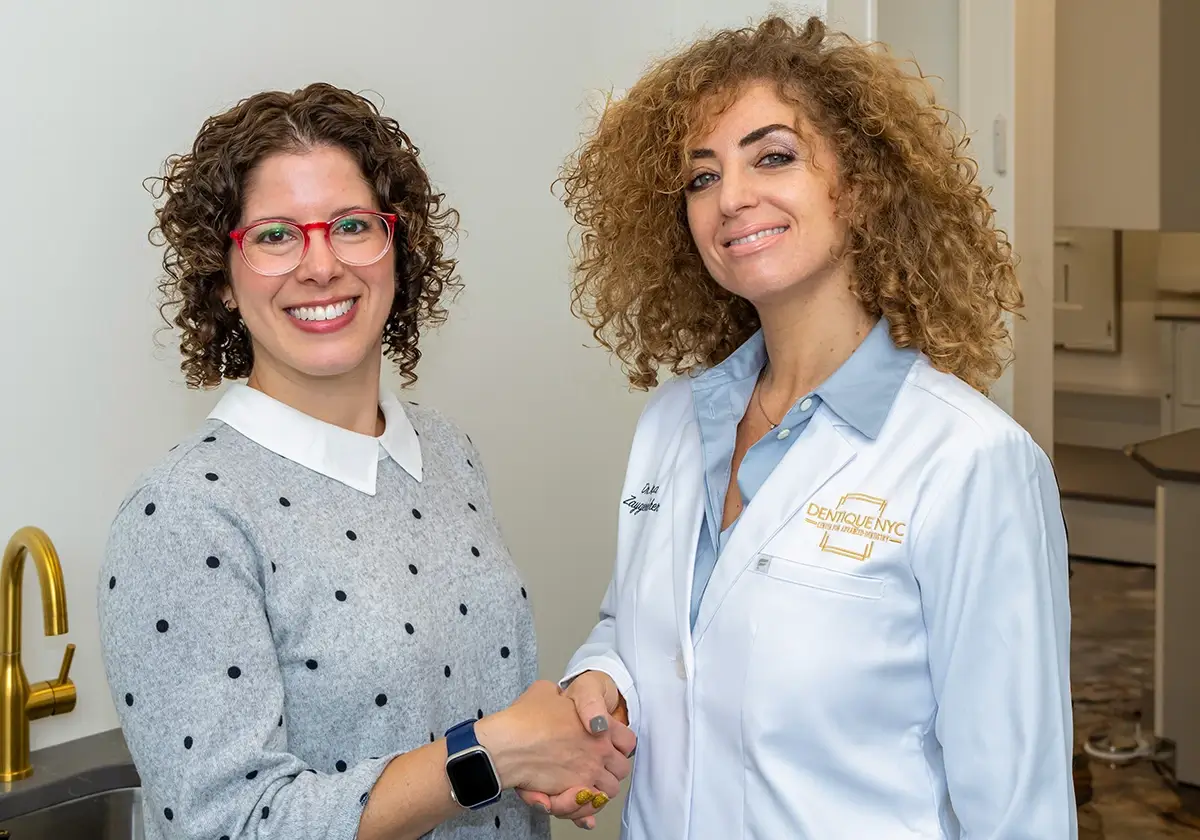 Two women smiling and shaking hands. The woman on the left has curly hair, red glasses, and is wearing a gray polka dot sweater. The woman on the right has curly hair and is wearing a white dental coat with a gold logo.