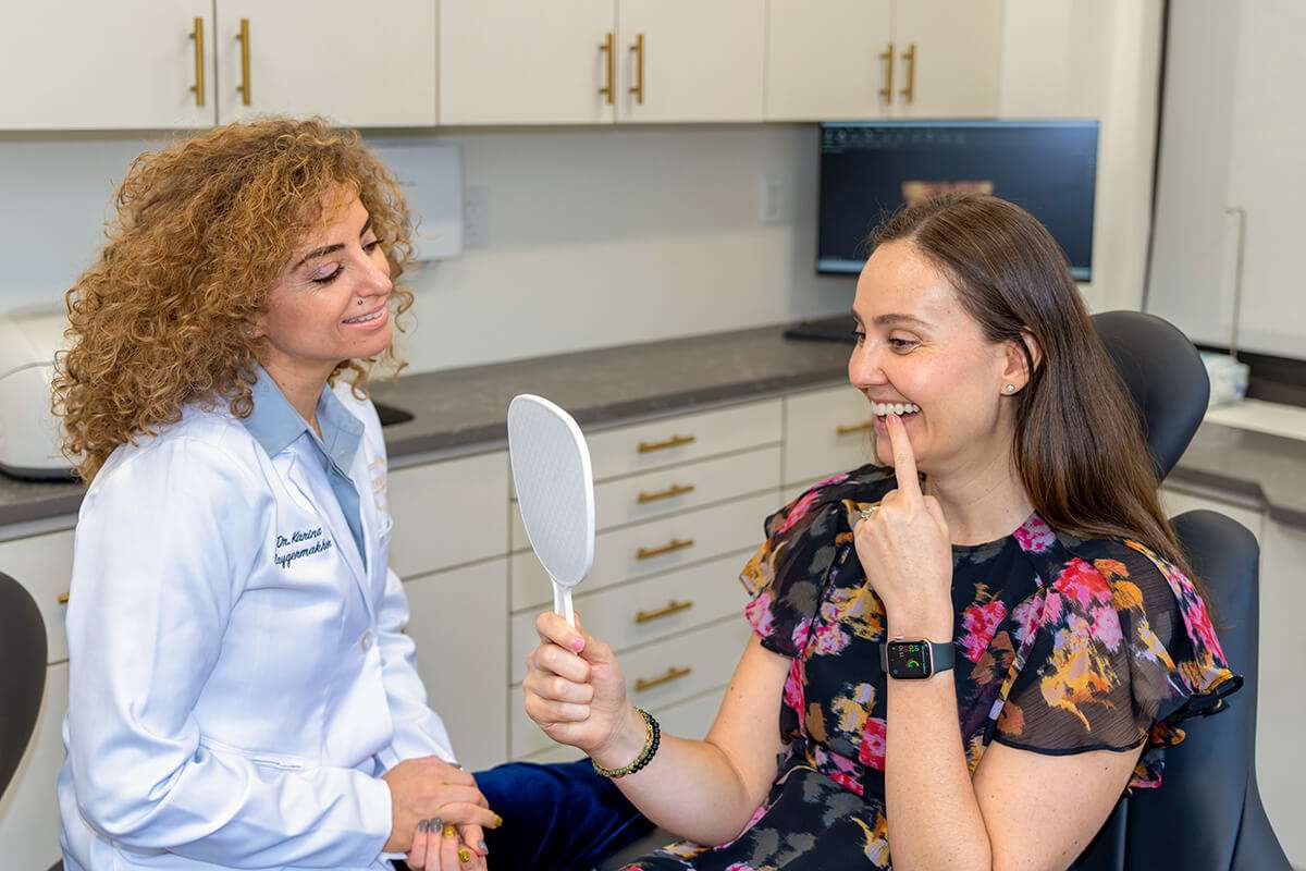 A dentist in a white coat talks to a smiling woman holding a mirror in a dental office. The woman points at her teeth, wearing a floral blouse and smartwatch. Background shows dental equipment and cabinetry.