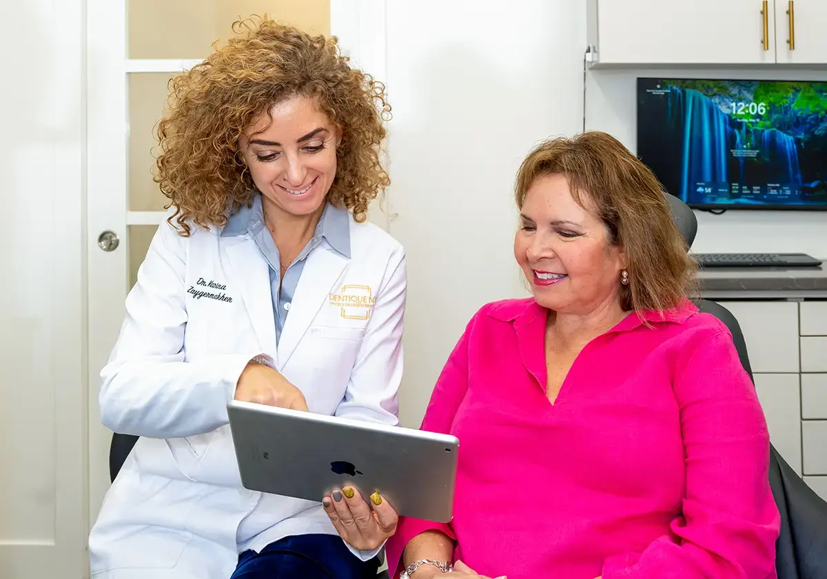 A dentist in a white coat shows a dental plan on a tablet to a smiling woman in a pink blouse. They are seated in a modern dental office with a monitor displaying a nature scene in the background.