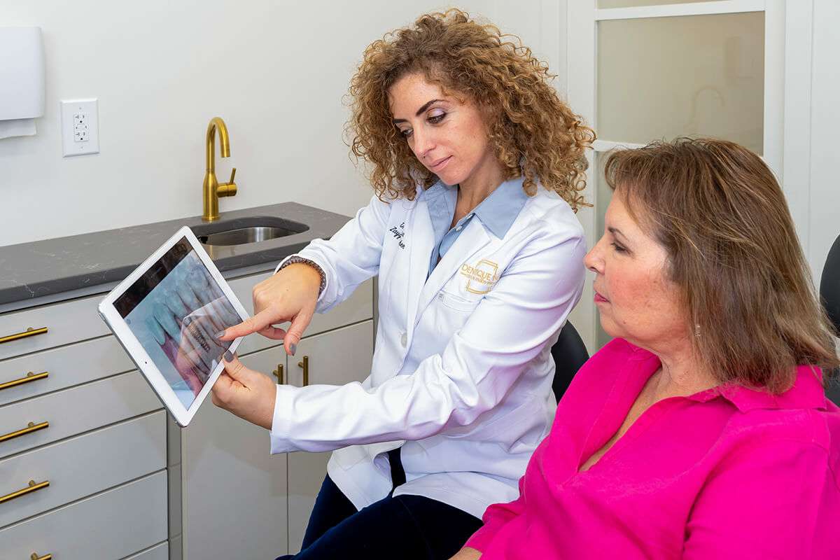 A dentist in a white coat shows a dental X-ray to a seated patient in a pink shirt. They are in a modern dental office with cabinetry and a sink in the background.