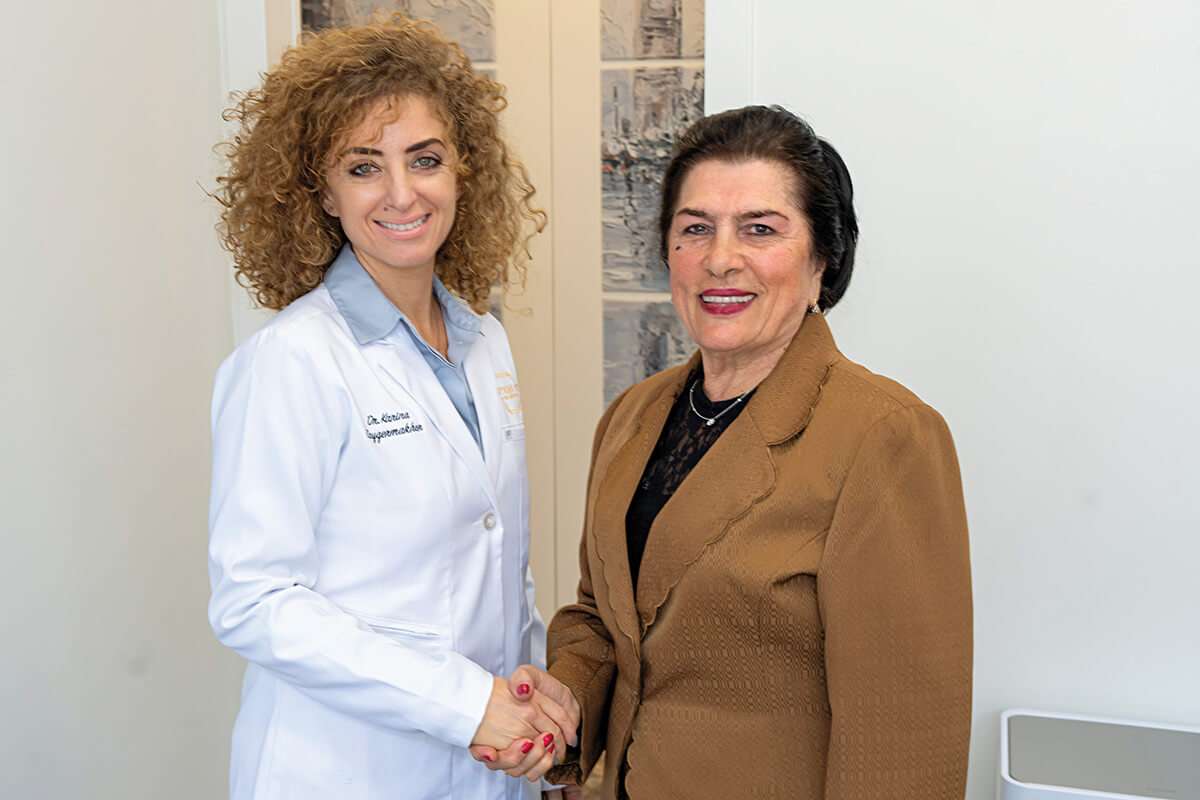 Two women smiling and shaking hands. One is wearing a white lab coat, suggesting she is a medical professional, while the other is in a brown blazer. They are standing in a bright room.