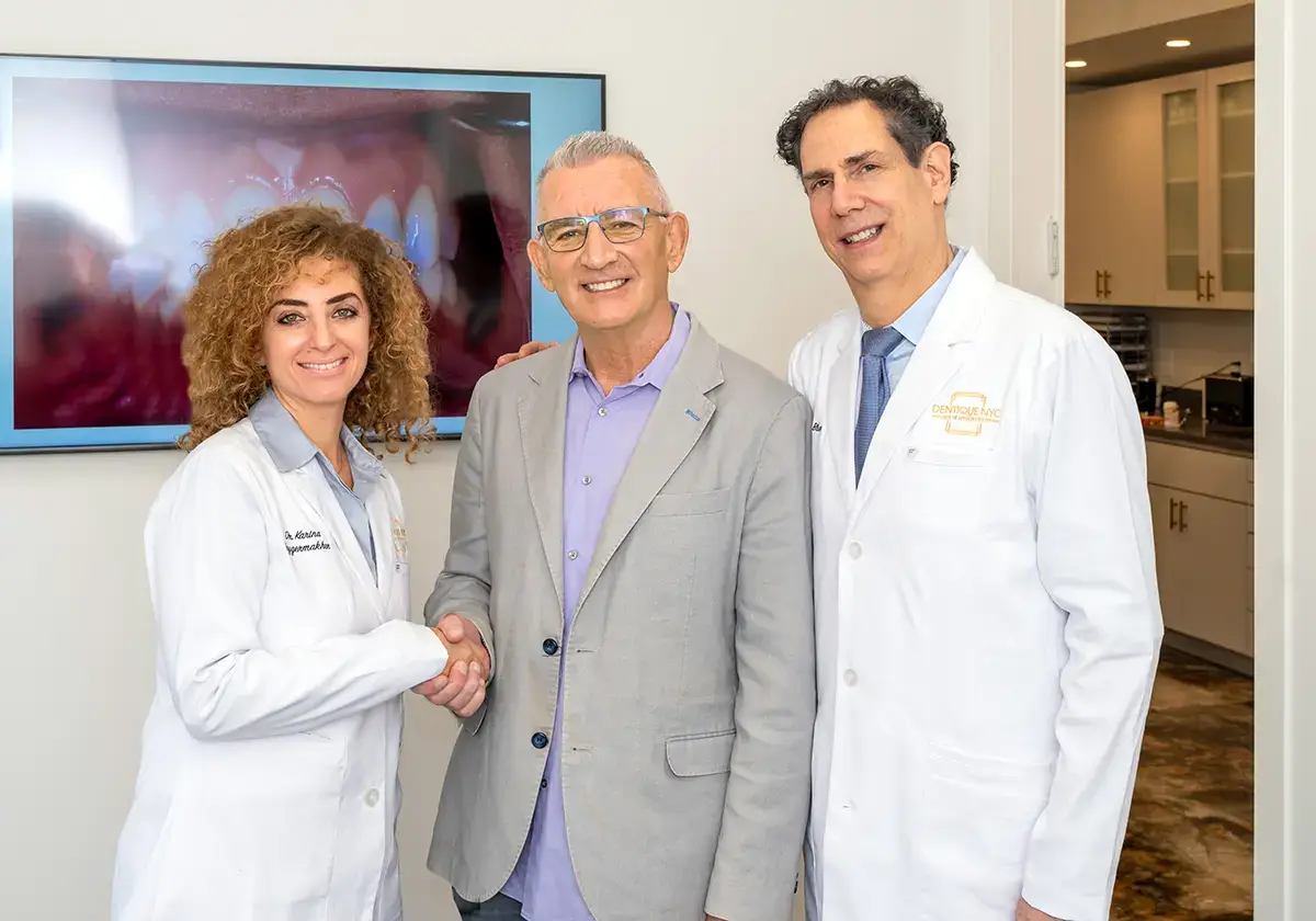 Two dentists in white coats smile while standing with a man in a gray blazer and purple shirt. The woman on the left shakes the mans hand. A monitor displaying a dental image is in the background.