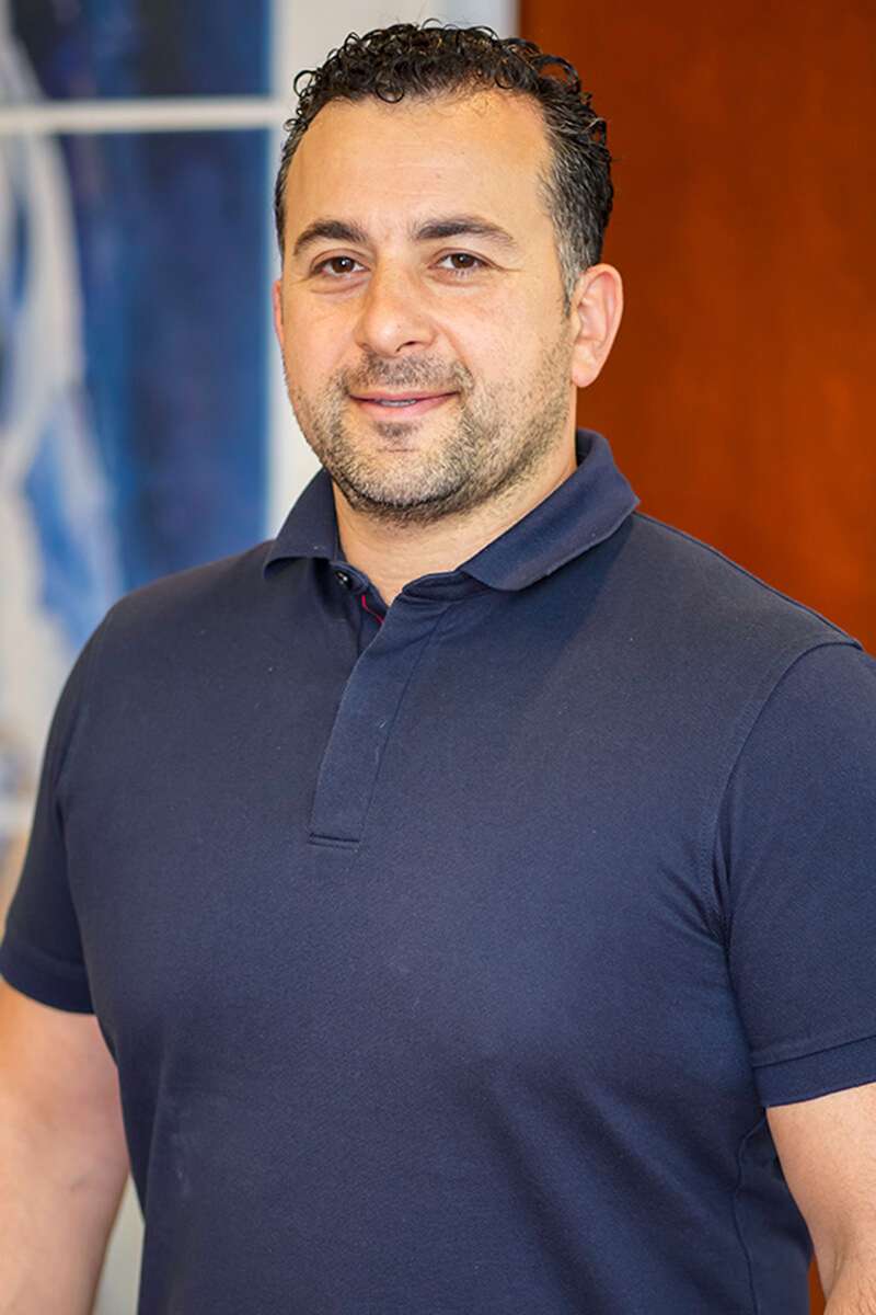 A person with short curly hair wearing a navy polo shirt is standing indoors, smiling slightly. The background features blurred artwork and a brown panel.