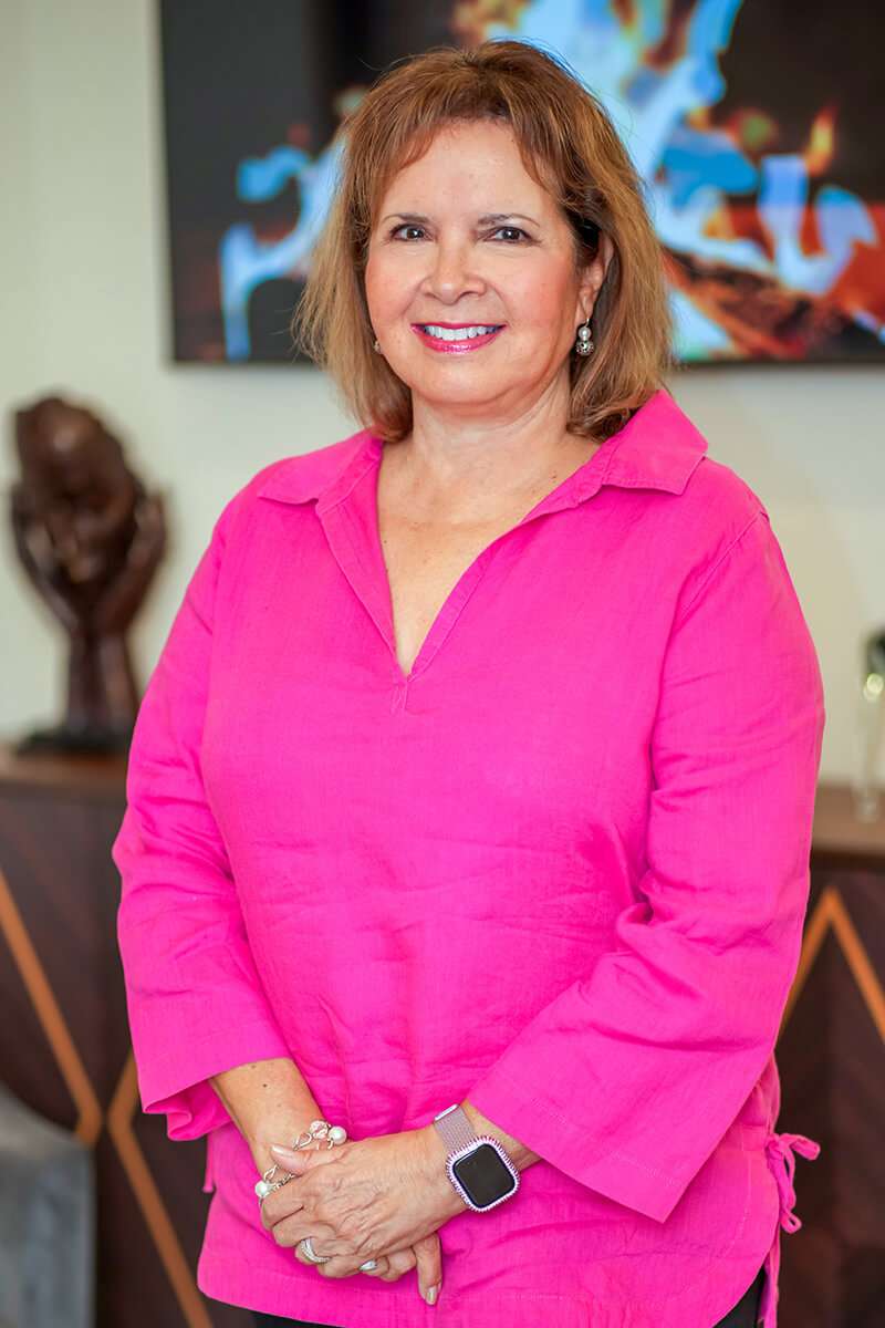 A woman in a bright pink blouse stands smiling. She has shoulder-length hair and wears a smartwatch and earrings. The background features abstract art and a sculpture, creating a warm, inviting setting.