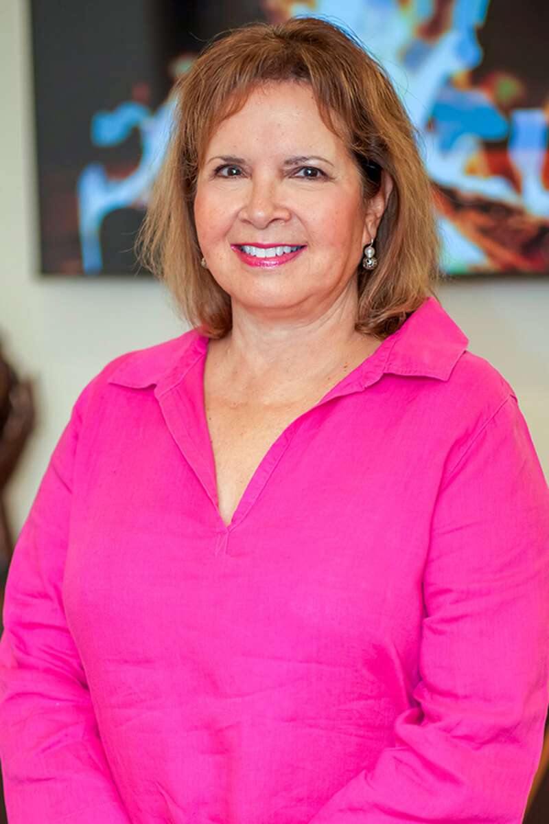 A woman with shoulder-length hair is smiling and wearing a bright pink blouse. She is standing indoors, and there is a blurred background with some colorful abstract shapes.