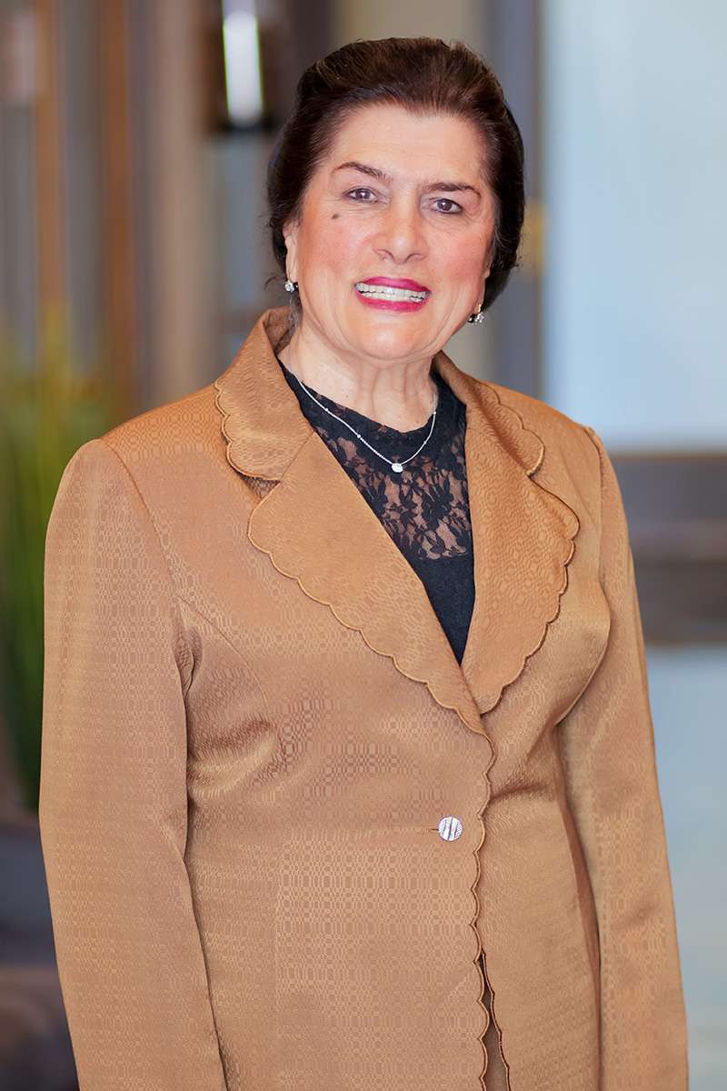 A woman in a brown blazer with a scalloped edge over a black lace top, standing and smiling against a blurred background.