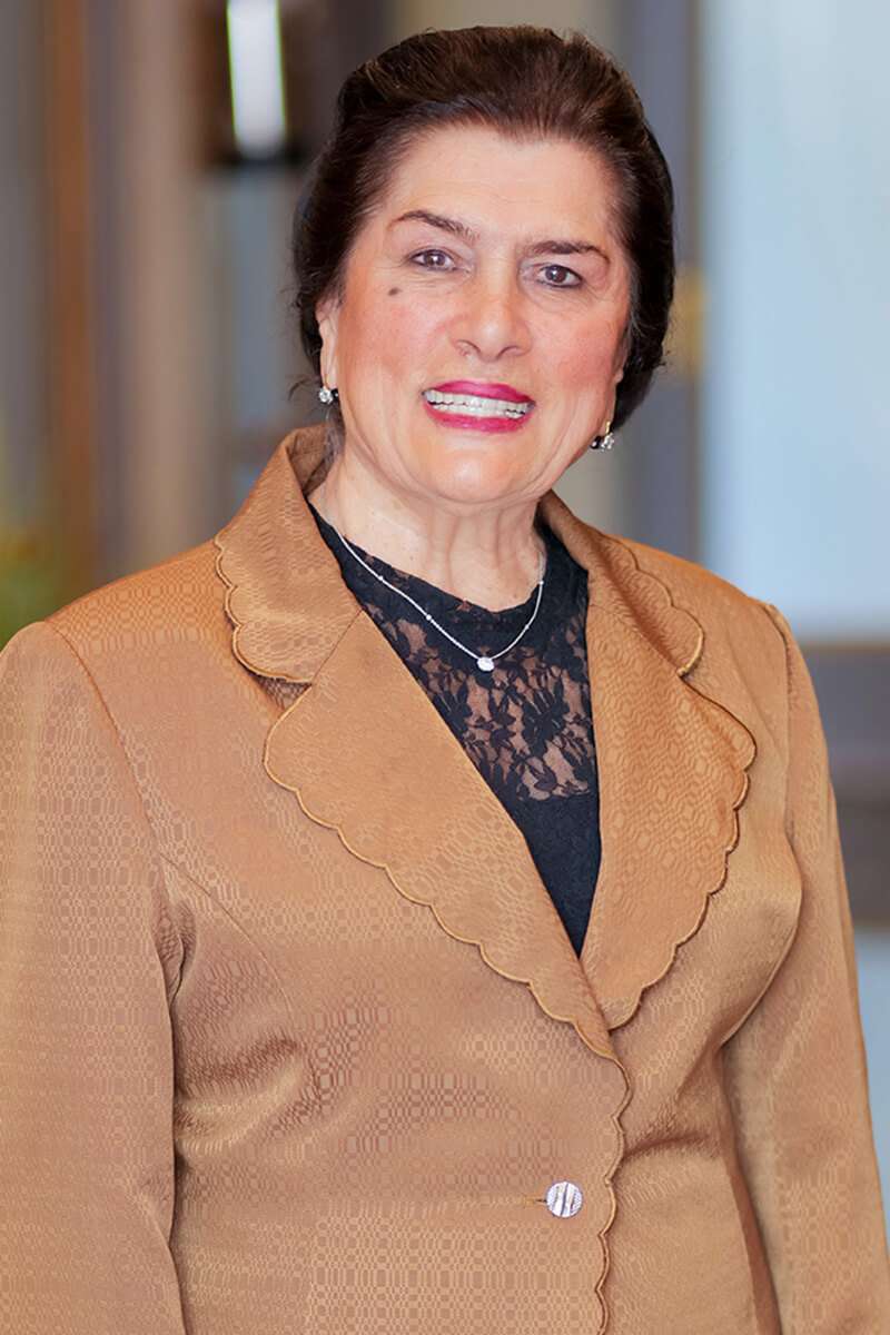 A smiling woman with dark hair, styled up, is wearing a brown blazer with a scalloped collar over a black lace top. She accessorizes with pearl earrings and a delicate necklace, standing against a softly blurred background.