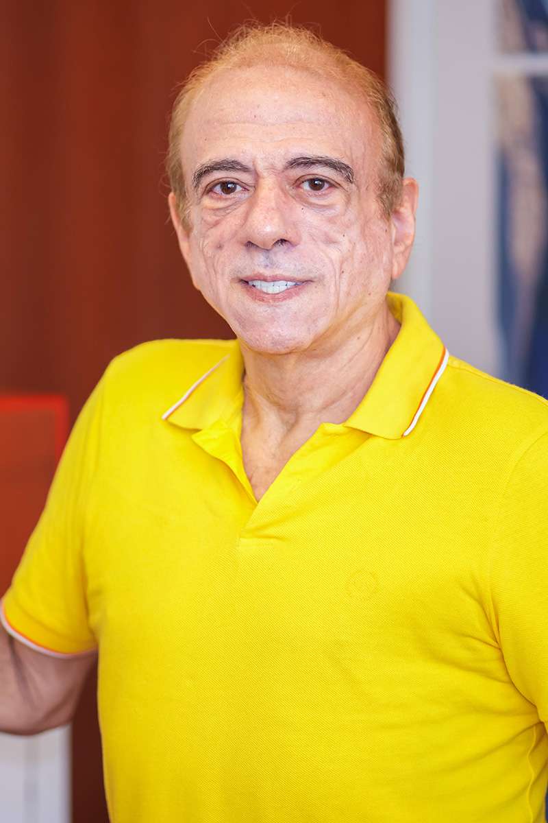 A person wearing a bright yellow shirt stands indoors, smiling. The background is softly blurred, featuring a mix of red and blue tones.