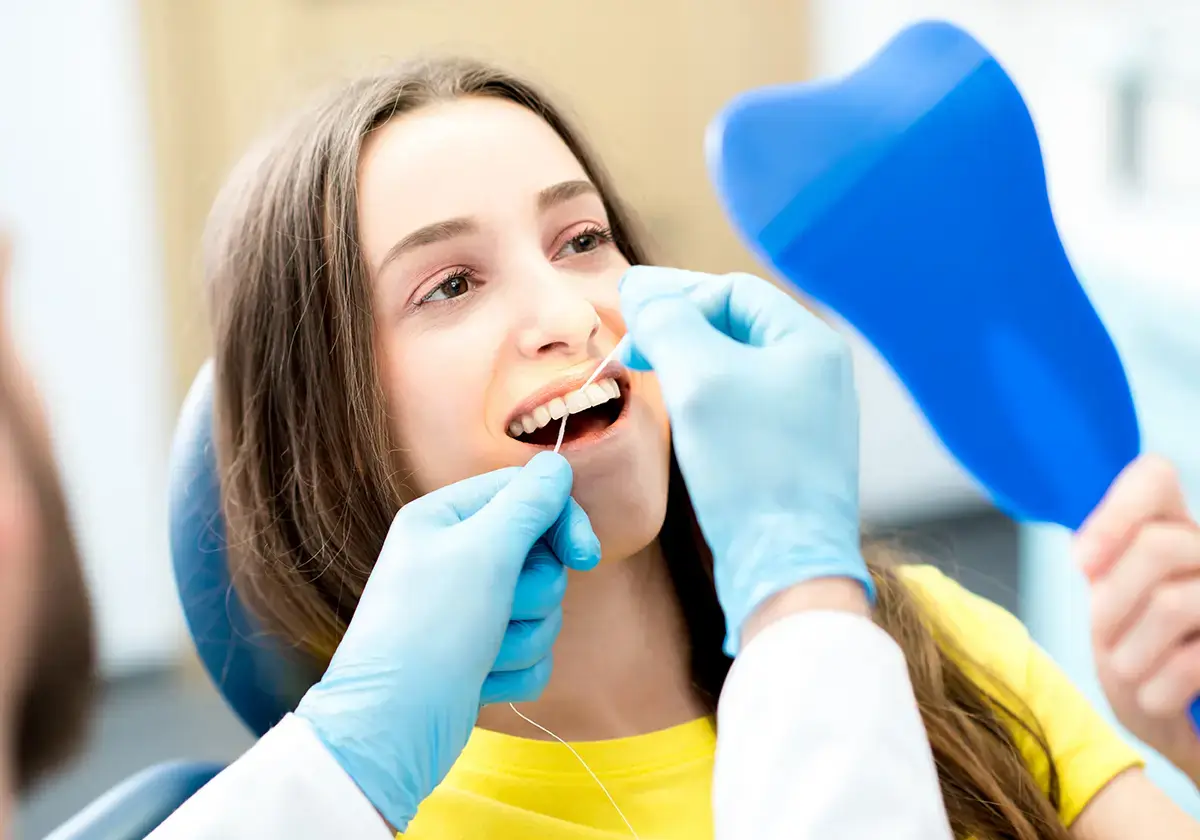 A young woman in a dental chair smiles while a dentist, wearing blue gloves, uses dental floss on her teeth. She holds a blue handheld mirror, observing the process.