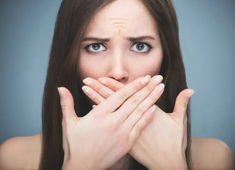 A woman with long brown hair looks directly at the camera, her eyebrows raised in concern. She covers her mouth with both hands, set against a plain gray background.